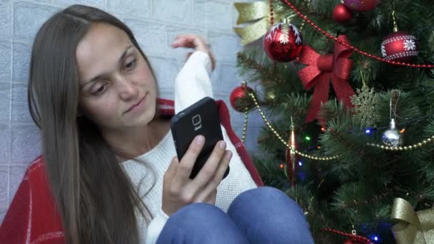 Frau sitzt mit rotem Karo und Smartphone auf dem Boden neben Weihnachtsbaum — Stockvideo