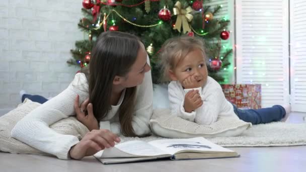 Moeder met dochter op de vloer liggen en het lezen van een boek in de buurt van de kerstboom — Stockvideo