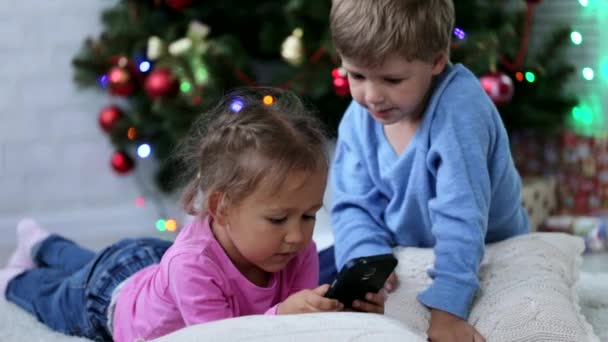 Lindos hermanos esperando algo divertido en el teléfono móvil cerca del árbol de Navidad . — Vídeos de Stock