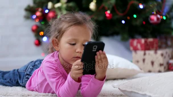 Kleines nettes Mädchen mit Smartphone vor dem Weihnachtsbaum. Nahaufnahme. — Stockvideo