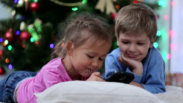 Nette Geschwister, die in der Nähe des Weihnachtsbaums irgendwas Lustiges im Handy wedeln. — Stockvideo
