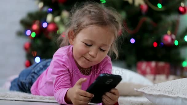 Niña linda usando el teléfono inteligente, delante del árbol de Navidad. Primer plano Shot . — Vídeo de stock