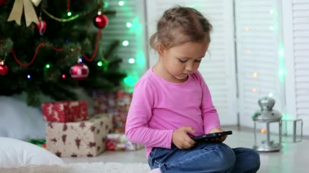 Niña linda usando el teléfono inteligente, delante del árbol de Navidad. Primer plano Shot . — Vídeo de stock