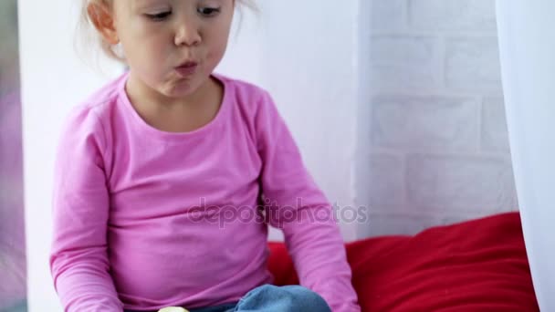 Портрет забавной маленькой девочки, сидящей на подоконнике и поедающей яблоко . — стоковое видео