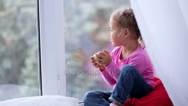 Retrato de niña linda divertida sentada en el alféizar de la ventana y comer manzana . — Vídeos de Stock