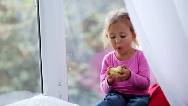 Porträtt av rolig söt liten flicka sitter på fönsterbrädan och äta äpple. — Stockvideo