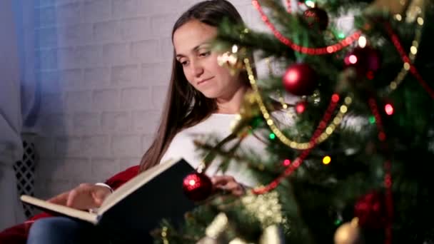 Schöne junge Frau sitzt auf einem Stuhl in der Nähe des Weihnachtsbaums und liest ein Buch. — Stockvideo