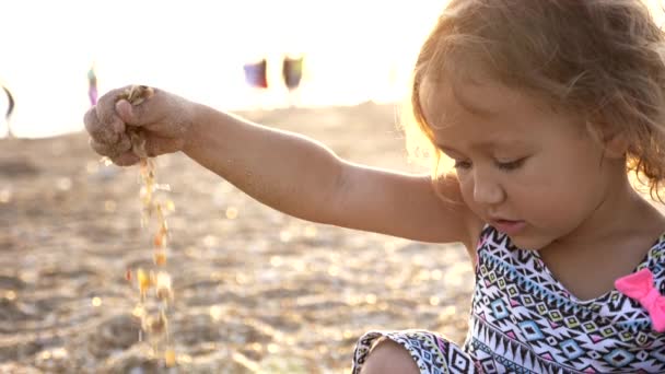 Маленькая девочка держит песок в руке на пляже и дает ему упасть снова . — стоковое видео