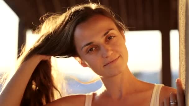 Молода красива жінка посміхається і дивиться на камеру на балконі на заході сонця . — стокове відео