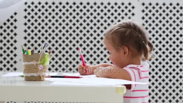 Симпатична дівчина дошкільного віку, що сидить за білим столом, зосереджена на малюванні чогось . — стокове відео