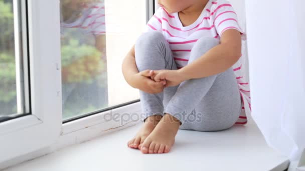 Gros plan des mains sur les genoux de mignonne petite fille assise sur le rebord de la fenêtre . — Video