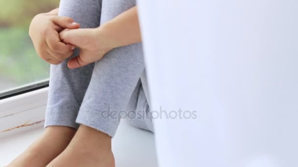 Primer plano de las manos sobre las rodillas de la linda niña sentada en el alféizar de la ventana de la ventana . — Vídeos de Stock