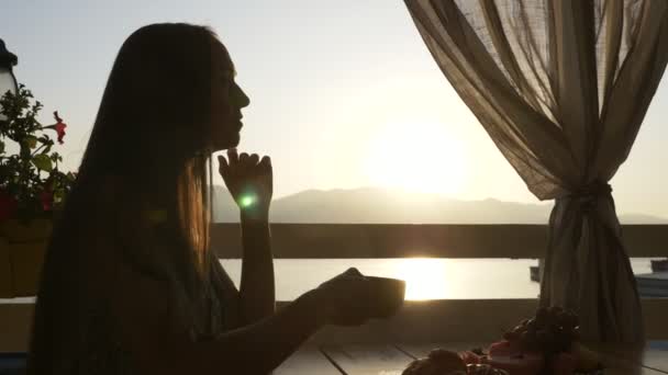 Silhouette of young woman sits in a cafe with panoramic views of sea bay. — Stock Video