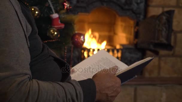 Senior mannen läser bok nära den öppna spisen och julgran på xmas eve — Stockvideo