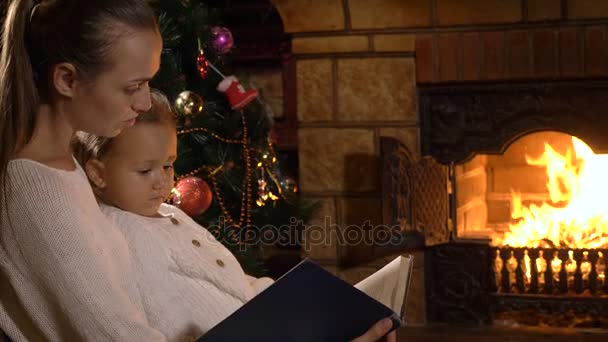 Madre con pequeña hija leyendo libro cerca de la chimenea y el árbol de Navidad — Vídeo de stock