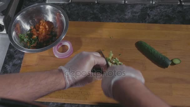 Professionele chef-kok in handschoenen snijden komkommer op houten bord op commerciële keuken — Stockvideo