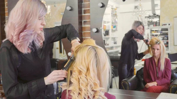 Woman hairdresser making curls at blond hair with curling irons at beauty salon.