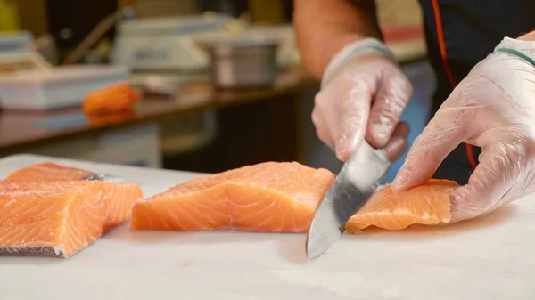 Close-up of sushi chef in gloves slices fresh salmon at sushi bar.