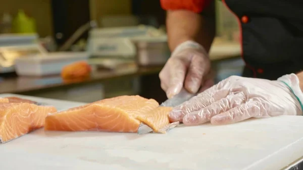Close-up of sushi chef in gloves slices fresh salmon at sushi bar.