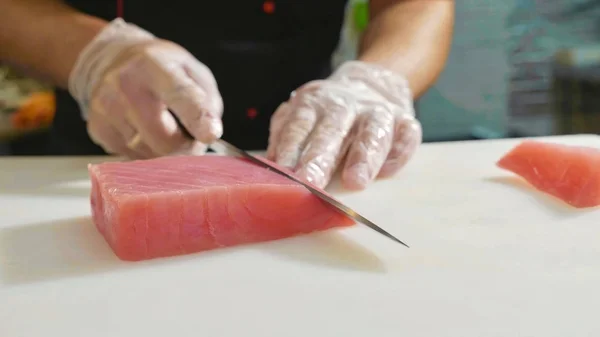 Close-up of sushi chef in gloves slices fresh red fish fillet at sushi bar.