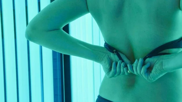 Young woman enters the tanning booth and removes a bra on the move.