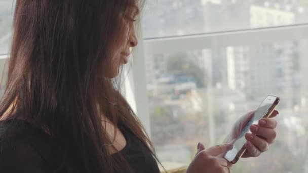 Hermosa chica sentada en la silla cerca de la ventana y el uso de teléfono inteligente — Vídeo de stock