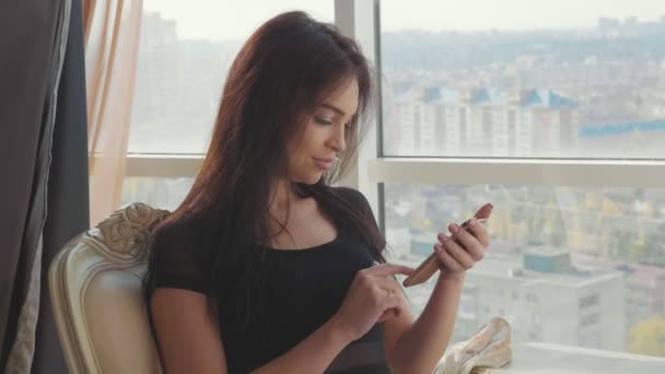 Piękna dziewczyna siedzi na krześle w pobliżu okna i przy użyciu smartfona — Wideo stockowe