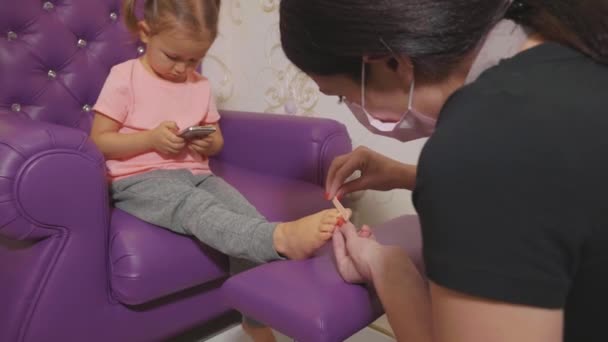 Bébé fille mignonne en utilisant un téléphone intelligent à la procédure de pédicure au salon de beauté spa . — Video