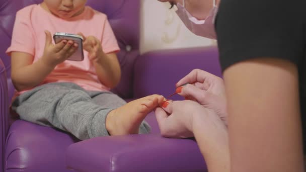 Bébé fille mignonne en utilisant un téléphone intelligent à la procédure de pédicure au salon de beauté spa . — Video