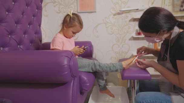 Cute dziewczynka za pomocą inteligentnego telefonu o pedicure procedury w salonie piękności spa. — Wideo stockowe