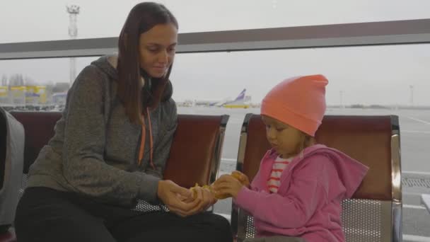Mother and daughter at airport. Little girl cleans mandarin at waiting hall. — Stock Video