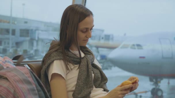 Jonge vrouw eet tangerine op luchthaven met vliegtuig op de achtergrond — Stockvideo
