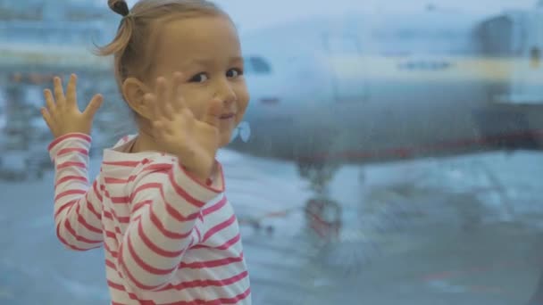 Kleines Mädchen steht am Flughafen am Fenster und winkt in die Kamera — Stockvideo