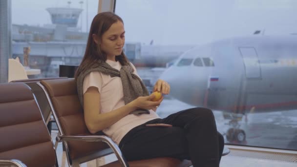 Молодая женщина ест мандарин в аэропорту с самолетом на заднем плане — стоковое видео