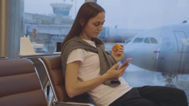 Fiatal nő eszik mandarin repülőtéren a repülőgép a háttérben — Stock videók