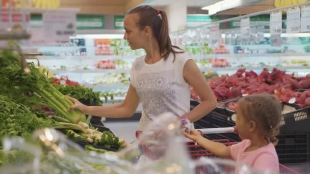 Jovem mãe com pequena filha escolhendo legumes verdes no supermercado — Vídeo de Stock