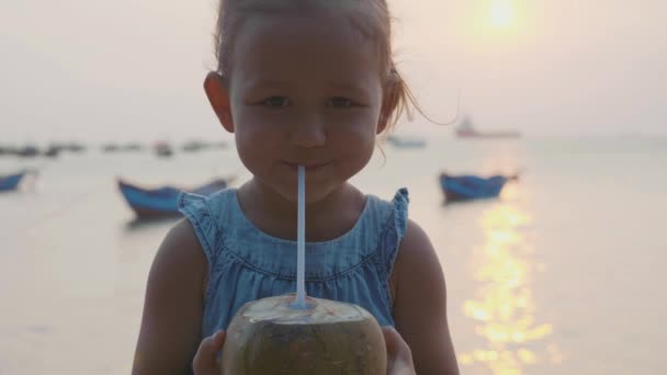 Little girl drinks coconut water at seafront on sunset in slow motion. — Stock Video