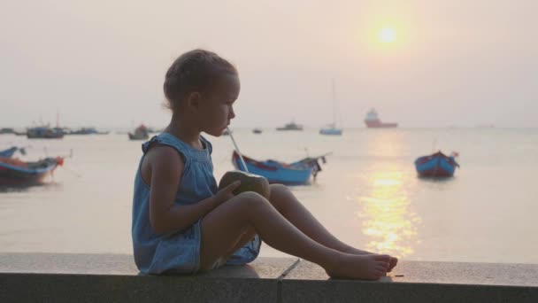 Lilla flickan dricker kokosvatten på havet på solnedgången i slow motion. — Stockvideo