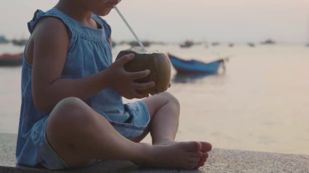 Malá holka pije kokosové vody u nábřeží na západ slunce v pomalém pohybu. — Stock video