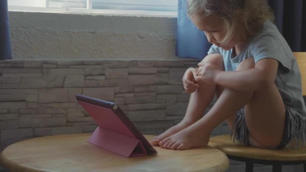 Pequeña niña linda sentada en la mesa y viendo algo en la tableta — Vídeos de Stock