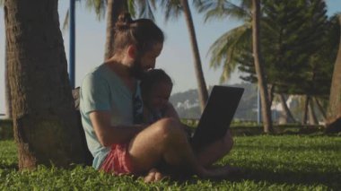 Doğa parkı ile laptop bir palmiye ağacının altında oturan adam.