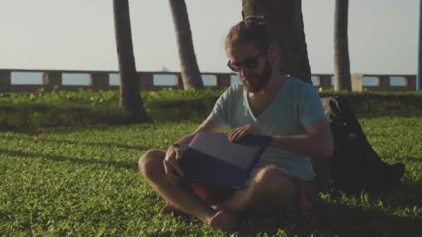 Man in nature park sitting with laptop under a palm tree. — Stock Video