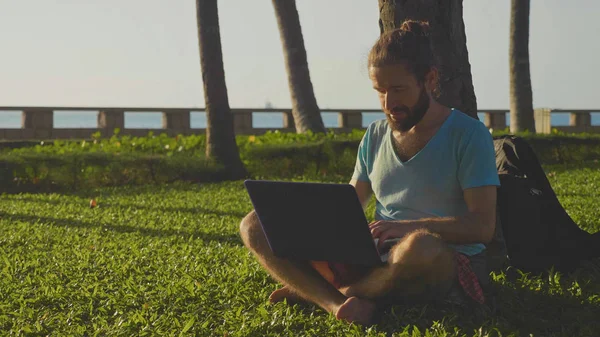 Man works in nature park. Male sitting with laptop under a palm tree — Stock Photo, Image