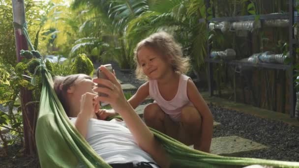 Молода жінка використовує смартфон у гамаку і грає зі своєю маленькою донькою — стокове відео