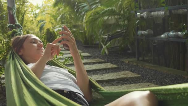 Mujer joven utiliza un teléfono inteligente mientras se encuentra en una hamaca en el jardín verde . — Vídeo de stock