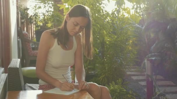 Mladá žena sedí u balkónu v slunečním světle a psaní deníku propisovací tužka — Stock video