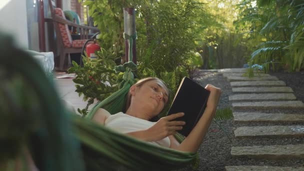 Junge Frau liest Buch liegend in Hängematte auf der Terrasse — Stockvideo