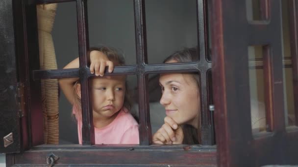 Madre e hija pequeña están mirando por la ventana con emociones aburridas . — Vídeos de Stock