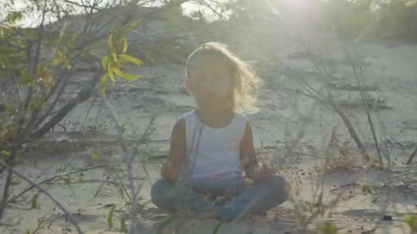 小さな女の子は、砂漠で瞑想します。日没の暖かい砂の上に座っている子. — ストック動画