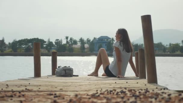 Молодая женщина путешественница сидит на деревянном пирсе и наслаждается природой в замедленной съемке — стоковое видео